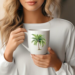May include: White ceramic mug with a green palm tree design. The mug is being held by a person.