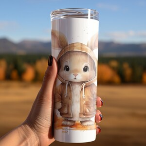 May include: A white stainless steel tumbler with a brown bunny wearing a brown hooded jacket. The bunny has brown eyes and a brown nose. The background is a blurred image of a forest.
