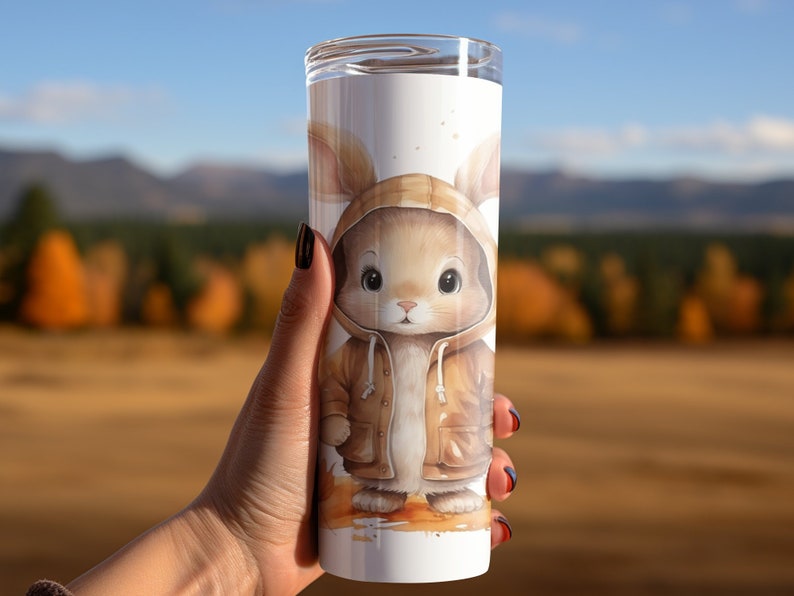 May include: A white stainless steel tumbler with a brown bunny wearing a brown hooded jacket. The bunny has brown eyes and a brown nose. The background is a blurred image of a forest.