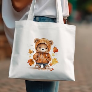 May include: White tote bag with a brown bear wearing a brown and orange jacket and standing on a bed of orange and yellow leaves.