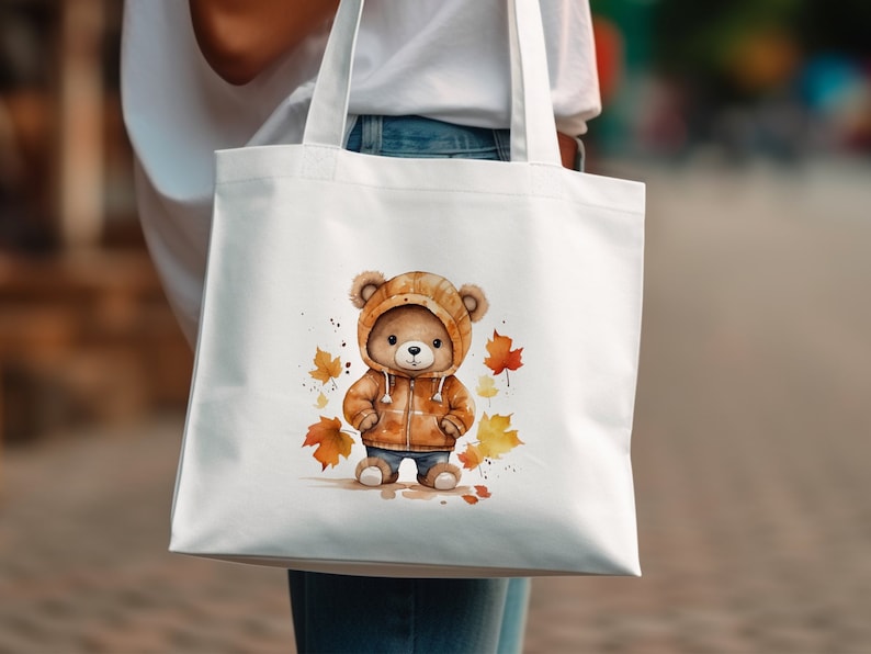May include: White tote bag with a brown bear wearing a brown and orange jacket and standing on a bed of orange and yellow leaves.