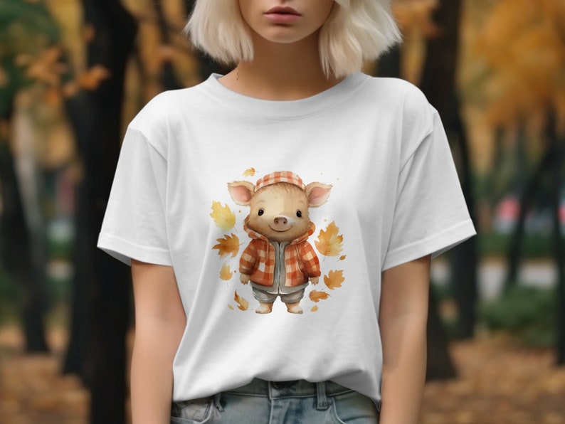 May include: White t-shirt with a cartoon pig wearing a red and black plaid jacket and a brown hat. The pig is surrounded by orange and brown leaves.