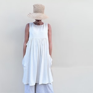 Puede incluir: Un vestido blanco sin mangas con cintura fruncida y dos bolsillos. El vestido se lleva con un sombrero de paja.