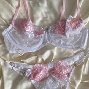 Könnte beinhalten: Weißes und rosafarbenes Spitzen-Dessous-Set mit Blumenstickerei. Der BH hat verstellbare Träger und ein zartes Blumenmuster. Das passende Höschen hat ein ähnliches Design und einen Satinbesatz.