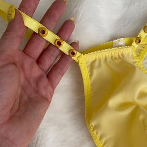 Könnte beinhalten: Hellgelbe Unterwäsche aus Netzstoff mit verstellbaren Trägern und goldfarbenen Metallösen. Das Kleidungsstück besteht aus einem halbtransparenten, fein gestrickten Stoff mit passender gelber Kante und einem gewellten Bund.