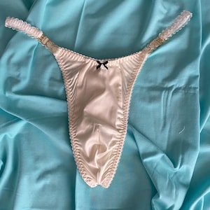 Könnte beinhalten: Ein weißer Satin-Tanga mit einer schwarzen Schleife und goldenen Verzierungen. Der Tanga hat verstellbare Träger mit einem weißen Spitzendetail.