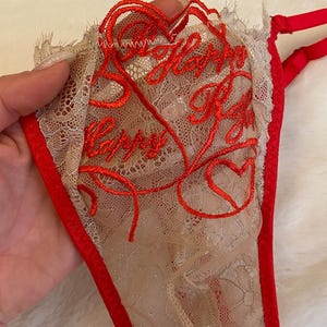 Könnte beinhalten: Beige Spitzen-String mit roter Bordüre und rotem gesticktem Text „Happy Valentine's Day“ mit Herz-Akzenten. Der String hat ein zartes, feminines Design.