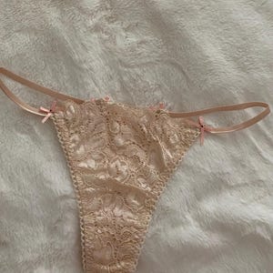 Könnte beinhalten: Ein beigefarbener Spitzen-Tanga mit zarten Blumenmustern und dünnen seitlichen Satinbändern. Das Kleidungsstück ist mit kleinen, hellrosa Schleifen an den Hüftansätzen und in der vorderen Mitte verziert, was dem Spitzenstoff ein Detail verleiht.