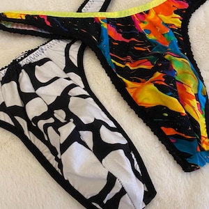 Könnte beinhalten: Zwei Unterhosen im String-Stil. Eine hat ein abstraktes Schwarz-Weiß-Muster, die andere ein lebendiges, farbenfrohes Spritzdesign mit neongelbem Besatz. Beide haben schwarze Verzierungen und liegen auf einer weißen, flauschigen Oberfläche.