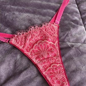 Puede incluir: Un tanga rosa brillante con intrincados detalles de encaje. El delicado patrón de encaje cubre la parte delantera y los bordes de la ropa interior. Las correas son de color rosa liso, sobre un fondo suave y texturizado.