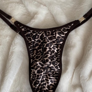 Pode incluir: Um fio dental com estampa de leopardo com alças marrons e detalhes decorativos dourados. A lingerie apresenta um padrão animal clássico em preto e bege. As alças são finas e ajustáveis, com pequenos enfeites decorativos.