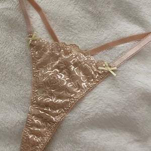 Könnte beinhalten: Ein beigefarbener Spitzen-Tanga mit Blumenmuster, dünnen elastischen Trägern und zwei kleinen hellgelben Schleifen an den Seiten, wo die Träger auf das vordere Spitzenteil treffen. Das Kleidungsstück ist mit zarten, gewellten Kanten versehen.