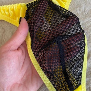 Puede incluir: Lencería amarilla con un panel de malla negra. Los bordes están ribeteados con tela amarilla. La lencería está hecha de un material suave y aterciopelado.