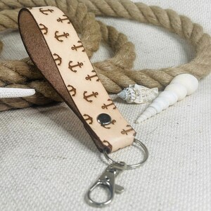 May include: A light brown leather keychain with a pattern of small anchors. The keychain has a silver metal key ring and a silver metal clasp.