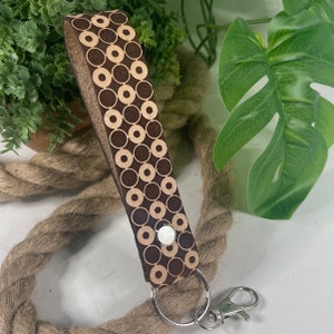May include: A brown leather keychain with a geometric pattern of beige circles. The keychain has a silver metal ring and a silver metal hook.