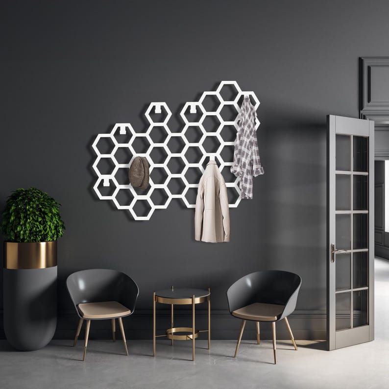 May include: White honeycomb-shaped wall mounted coat rack with hooks. The rack is mounted on a dark gray wall. Two chairs and a small table are in the foreground.