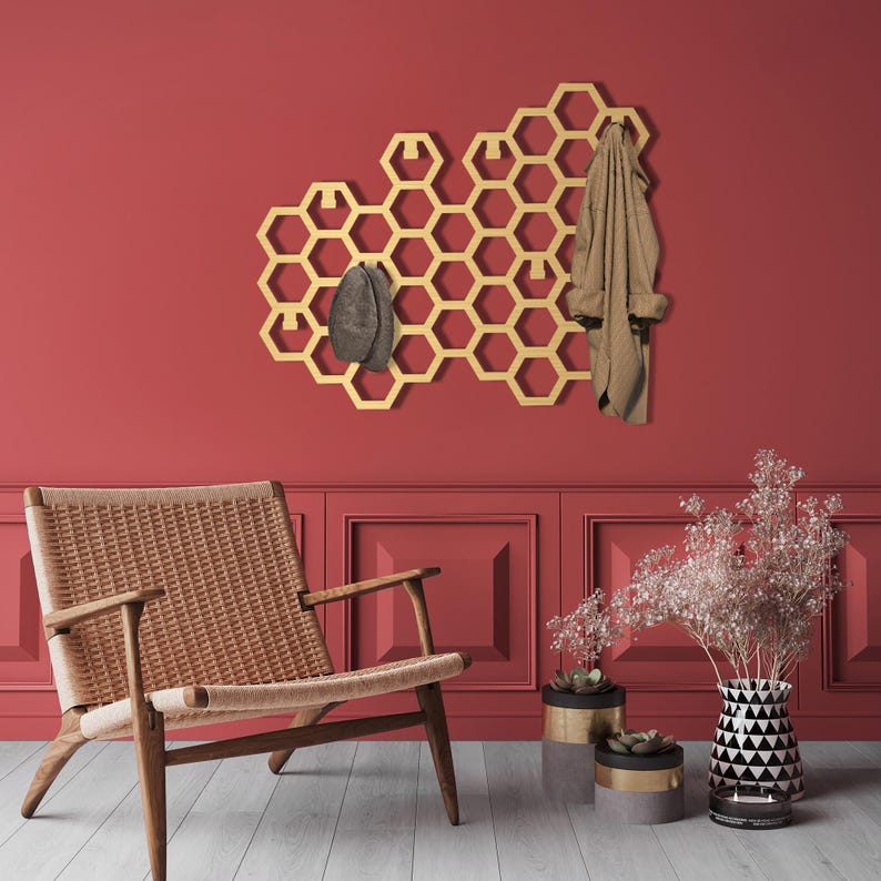 May include: Gold honeycomb wall mounted coat rack with hooks, a brown hat, and a brown jacket hanging on it. The rack is mounted on a red wall with a brown wicker chair and potted plants in front of it.