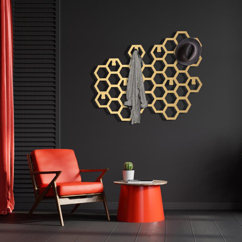 May include: A gold honeycomb-shaped wall mounted coat rack with a hat and scarf hanging on it. The rack is mounted on a black wall. A red armchair and a red side table are in the foreground.