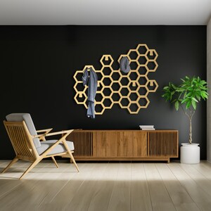 May include: A gold honeycomb-shaped wall mounted coat rack with hooks. The rack is mounted on a black wall and has a wooden shelf below it. A chair and a potted plant are in the foreground.