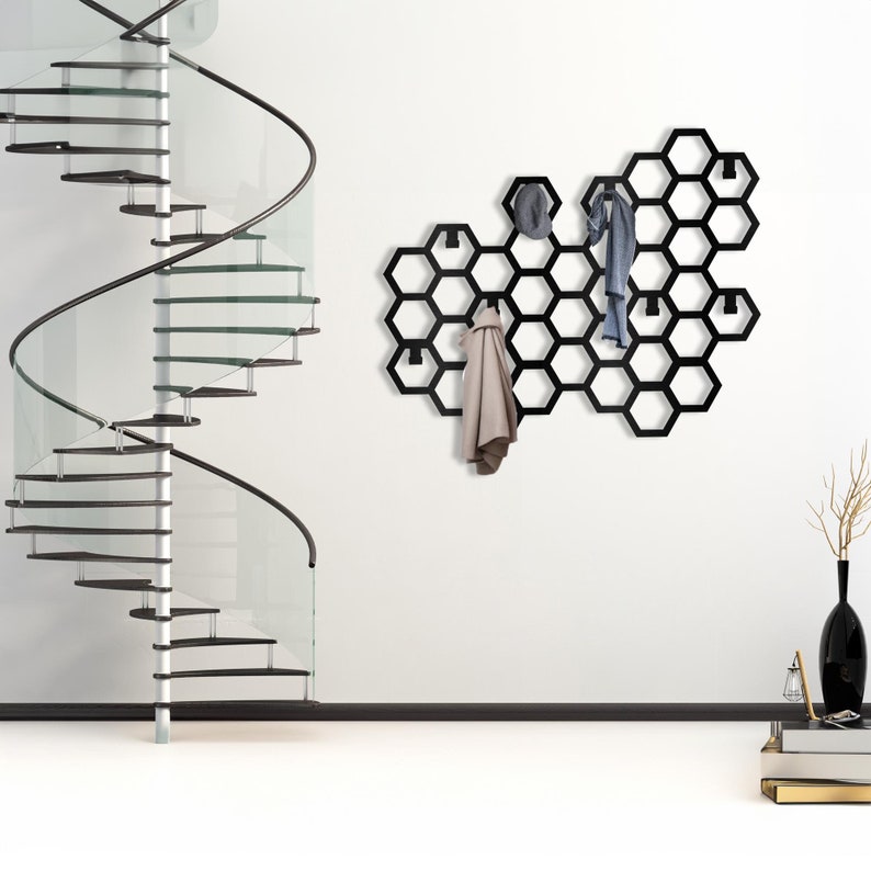 May include: Black metal hexagonal wall-mounted coat rack with hooks. The rack is shaped like a honeycomb and has a modern design. It is mounted on a white wall and has a scarf and jacket hanging from it.