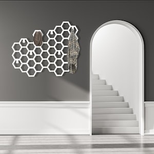 May include: White honeycomb-shaped wall mounted coat rack with a plaid shirt and a brown hat hanging on it. The rack is mounted on a gray wall with white trim. A white staircase is visible in the background.