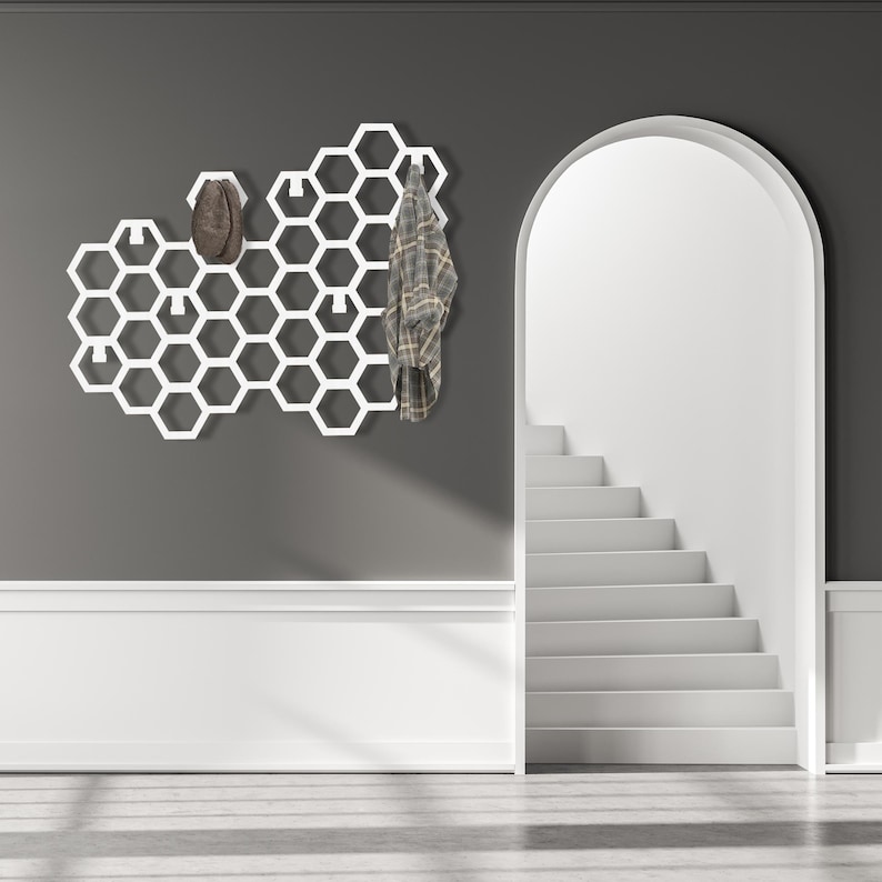 May include: White honeycomb-shaped wall mounted coat rack with a plaid shirt and a brown hat hanging on it. The rack is mounted on a gray wall with white trim. A white staircase is visible in the background.