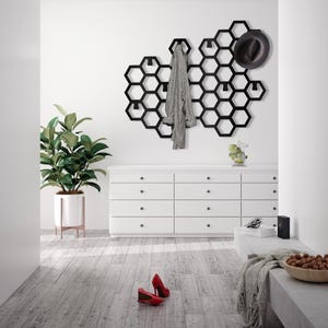 May include: A black metal honeycomb-shaped wall organizer with hooks for hanging coats, hats, and other items. The organizer is mounted on a white wall above a white dresser with six drawers. A gray sweater is hanging on one of the hooks, and a black hat is hanging on another hook.