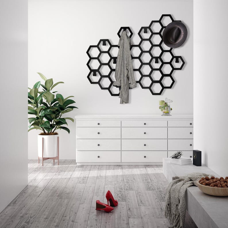 May include: A black metal honeycomb-shaped wall organizer with hooks for hanging coats, hats, and other items. The organizer is mounted on a white wall above a white dresser with six drawers. A gray sweater is hanging on one of the hooks, and a black hat is hanging on another hook.