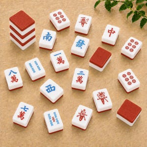 May include: A collection of rectangular Mahjong tiles featuring white faces with red and blue Chinese characters, numbers, and symbols. Several tiles are displayed face-up, while others are stacked or placed face-down showing red textured backs.