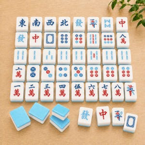 May include: A collection of white mahjong tiles with blue and red engraved characters, symbols, and numbers. The set includes a grid of upright tiles and several loose tiles placed face-up and face-down, displaying various suits and honors.