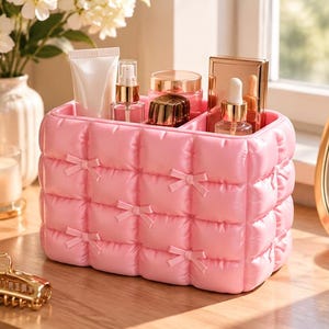 Op de afbeelding: Een rechthoekige roze cosmetica-organizer met een gewatteerde textuur en kleine strikjes op elk kruispunt. De houder heeft meerdere binnenvakken met diverse huidverzorgingsflesjes, een tube en een goudkleurige compacte spiegel.