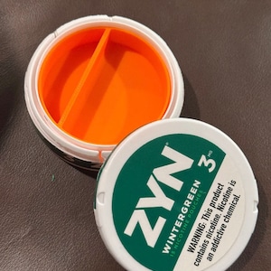 May include: A round white plastic container with an open top revealing a bright orange interior divided into two sections. The detached white lid features a green circular label with the text ZYN WINTERGREEN 3 MG 15 NICOTINE POUCHES.