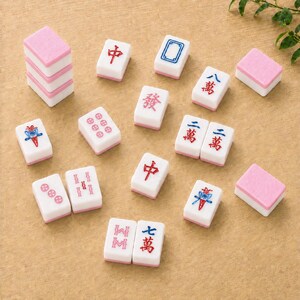 May include: A collection of rectangular Mahjong tiles featuring white faces with red, blue, and green engraved symbols and characters, paired with solid pink backs. Some tiles are stacked, while others are scattered, displaying various game markings.