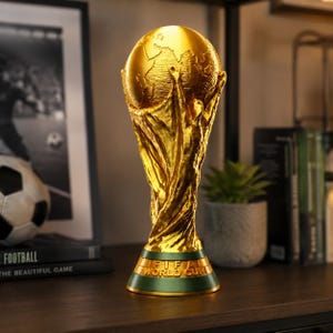 May include: A gold-colored trophy featuring a globe supported by two stylized figures with raised arms. The base is green with the text FIFA WORLD CUP embossed in gold lettering. The design depicts a dynamic, textured surface representing the world.