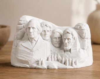May include: A white matte sculpture depicting the Mount Rushmore National Memorial. The piece features the carved faces of four United States presidents set against a rocky mountain backdrop with a small row of pine trees at the base.