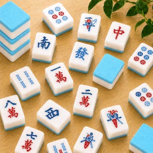 May include: A collection of white Mahjong tiles with blue and red engraved characters, symbols, and floral designs, including some plain blue-backed tiles, scattered across a light wood surface with a small green plant visible in the corner.