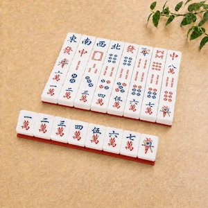 May include: A set of white rectangular Mahjong tiles featuring red and blue engraved Chinese characters and symbols. The tiles are arranged in a neat grid and a single horizontal row, displaying various numerical values and directional markers.
