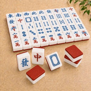 May include: A set of white Mahjong tiles with red and blue engraved characters, symbols, and numbers. The tiles are arranged in a rectangular grid with several loose tiles placed in the foreground, showing both the front faces and red backs.