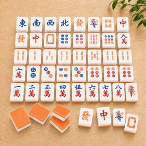 May include: A collection of white rectangular Mahjong tiles with orange backs, featuring various engraved Chinese characters, numbers, and circular patterns in blue, red, and green ink, arranged in a grid with several loose tiles at the bottom.