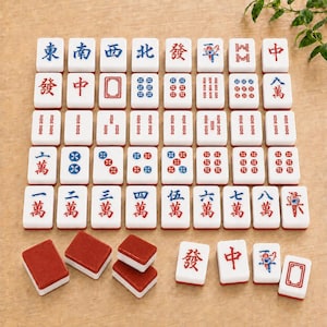 May include: A set of white rectangular mahjong tiles featuring various red, blue, and black engraved Chinese characters, symbols, and numerical patterns. Some tiles are displayed face-up in a grid, while others are placed face-down showing red backs.