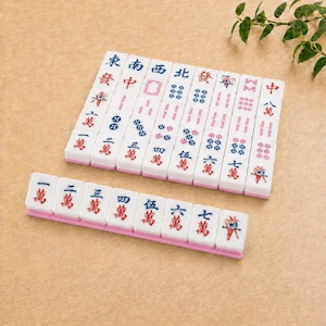 May include: A set of white rectangular mahjong tiles with pink bases, featuring various blue and red Chinese characters, numerical symbols, and decorative icons. The tiles are arranged in a neat grid and a single horizontal row on a tan surface.