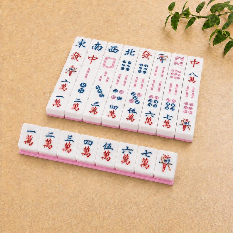 May include: A set of white rectangular mahjong tiles with pink bases, featuring various blue and red Chinese characters, numerical symbols, and decorative icons. The tiles are arranged in a neat grid and a single horizontal row on a tan surface.