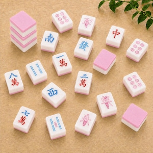 May include: A collection of rectangular mahjong tiles with white faces and pink backs. The tiles feature various engraved symbols, characters, and numbers in red, blue, and pink ink. Some tiles are stacked, while others are scattered across the surface.