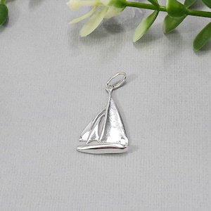 Könnte beinhalten: Ein silberner Segelboot-Anhänger. Der Anhänger ist ein schlichtes, minimalistisches Design mit einer glatten, polierten Oberfläche.
