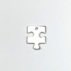 Może przedstawiać: Srebrny charm w kształcie puzzla z otworem na górze do przymocowania do naszyjnika lub bransoletki.