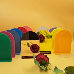 Op de afbeelding: Een verzameling kleurrijke, gebogen acryl display stands. De stands zijn in verschillende kleuren, waaronder groen, roze, blauw, rood, zwart, geel en oranje. Een rode roos is voor de stands geplaatst, met een gele achtergrond.