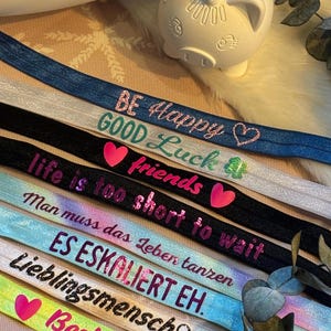 Könnte beinhalten: Ein Stapel bunter Gummibänder für Haare mit verschiedenen inspirierenden Botschaften darauf gedruckt. Die Botschaften lauten: "Be Happy", "Good Luck", "Friends", "Life is too short to wait", "Man muss das Leben tanzen", "ES ESKALIERT EH", "Lieblingsmensch" und "Besties".