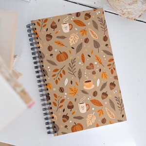 Op de afbeelding: Spiraalgebonden notitieboek met een bruinbeige kaft met een herfstthema. Het ontwerp bevat pompoenen, koffiekopjes, eikels, bladeren en koffiebonen in bruin, oranje en wit. Het notitieboek is open.