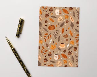 Puede incluir: Un cuaderno con un estampado de temática otoñal con calabazas, bellotas, tazas de café y hojas en tonos marrones, naranjas y beige. Un bolígrafo negro y dorado está al lado del cuaderno.
