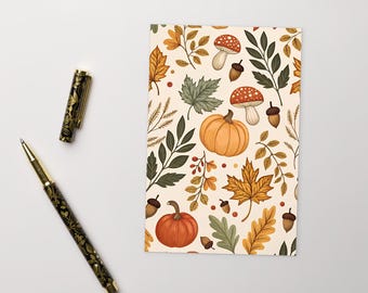 Puede incluir: Un bolígrafo decorativo con un diseño floral negro y dorado junto a un cuaderno con un patrón de temática otoñal. El cuaderno presenta calabazas, hojas, bellotas y setas en tonos naranja, verde y marrón sobre un fondo crema.